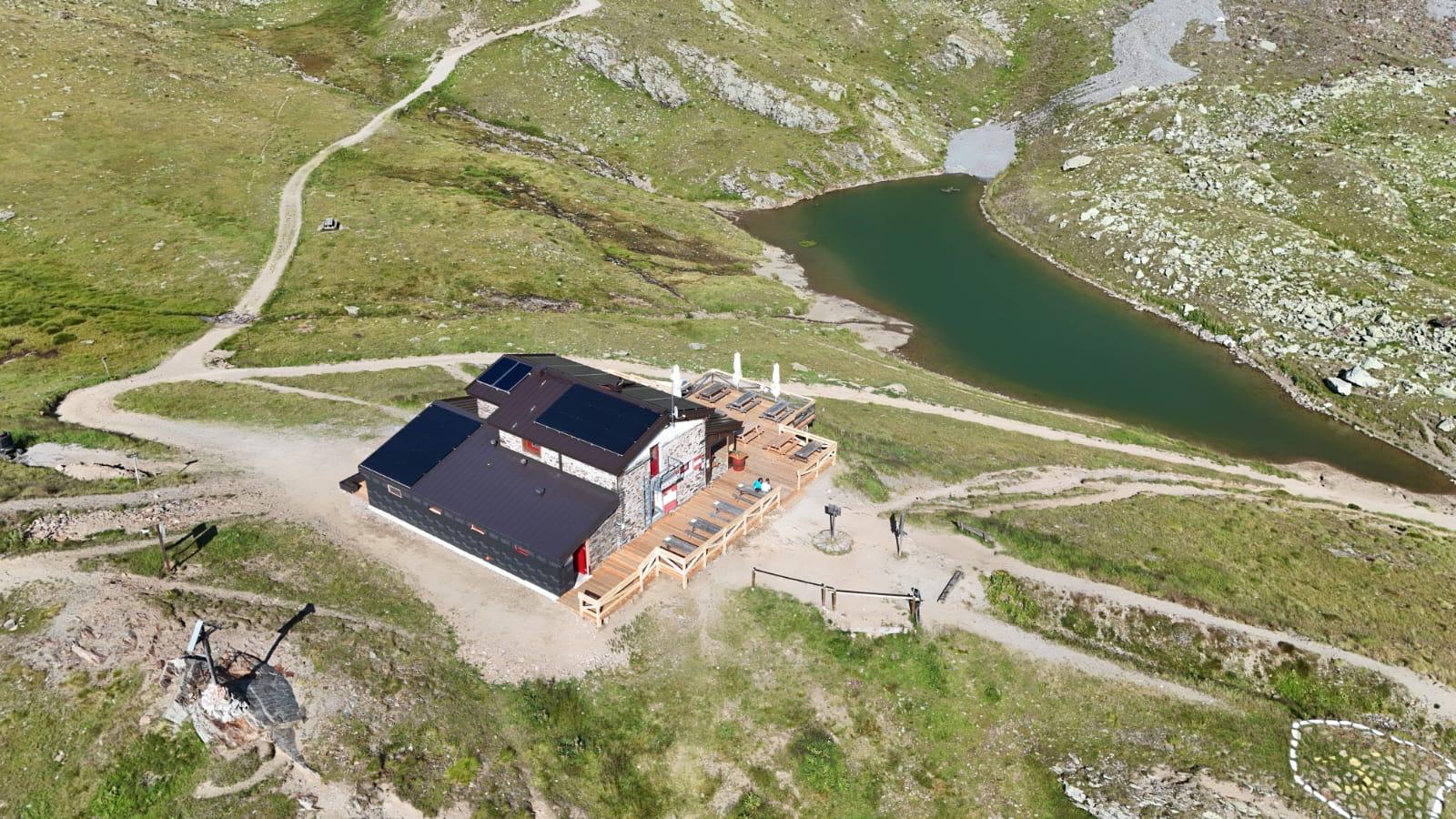 Veduta aerea del Rifugio Bozzi: l'edificio in pietra con la terrazza in legno e gli ombrelloni, un laghetto alle spalle e i sentieri che convergono nella conca